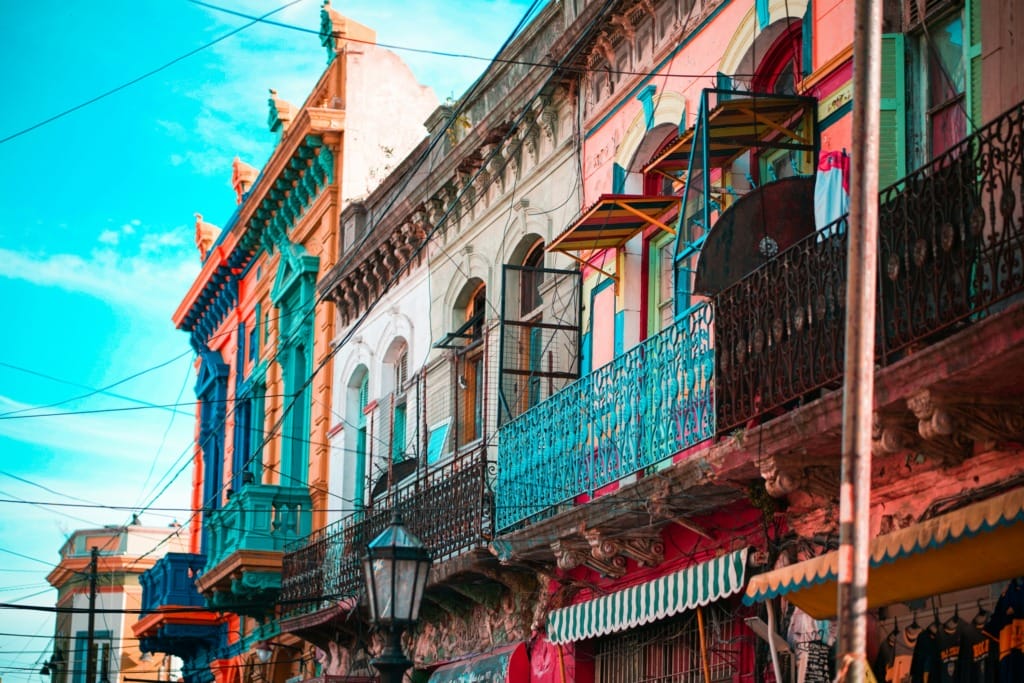 Buenos Aires'in rengarenk mahallelerinden, özellikle de La Boca'nın canlı ruhunu yansıtan bu görselde, balkonları ve demir parmaklıkları turkuaz, pembe ve sarı gibi göz alıcı tonlarda boyanmış eski mimariye sahip binalar, gökyüzünde kesişen sayısız elektrik teli ve karakteristik şemsiyelerle tutku dolu bir atmosfer sunuyor.