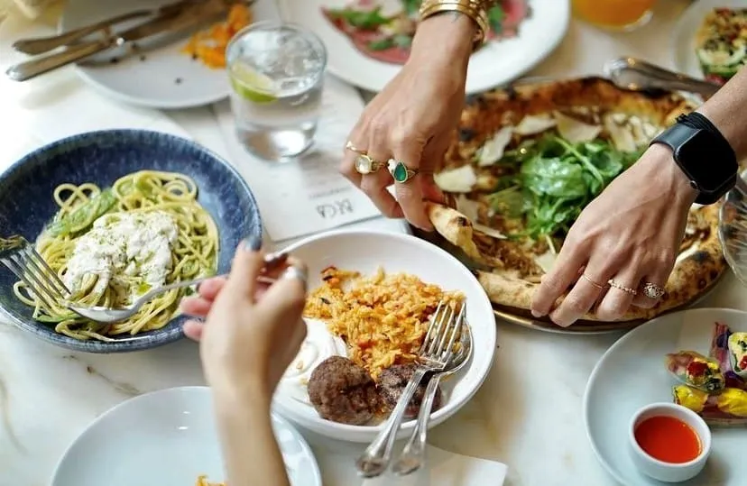 Beca İstanbul restoranında, bir masada pesto soslu spagetti, bol rokalı ve parmesan dilimli pizza, pilav ve köftenin yanı sıra çeşitli başlangıçların olduğu zengin bir yemek deneyimi yaşanırken, insanlar keyifle yemeklerini paylaşıyorlar.