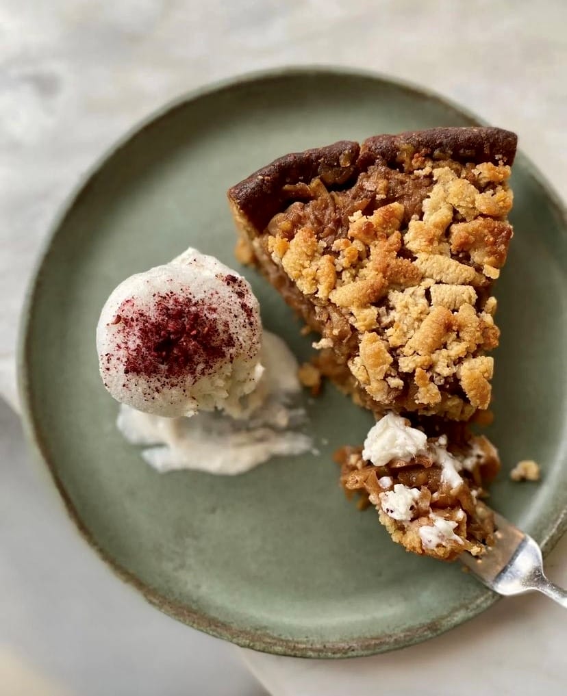 Vegan crumble dilimi, üzerinde kırmızı toz bulunan bitki bazlı dondurma topuyla birlikte rustik yeşil bir tabakta sunulurken, çatalla alınmış bir lokması ile Etiler'in en iyi mekanları arasında yer alan Bi Nevi Deli'nin yaratıcı ve bitki bazlı tatlılarını vurguluyor.