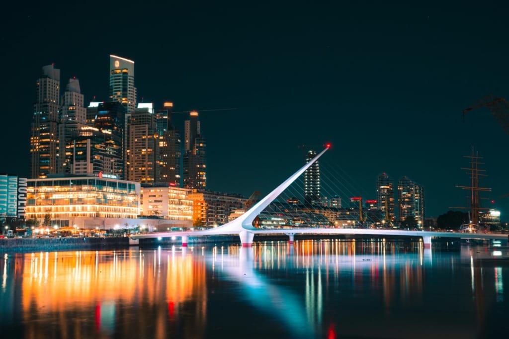 Buenos Aires'in modern yüzü olan Puerto Madero bölgesindeki ikonik Puente de la Mujer köprüsü, gece vakti şehrin ışıltılı yüksek binalarının yansımalarıyla sakin nehri turuncu, mavi ve beyaz tonlarında büyüleyici bir şekilde aydınlatarak şehrin tutkulu ve dinamik atmosferini gözler önüne seriyor.