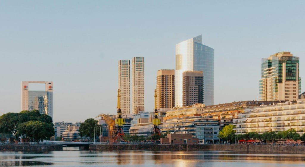 Gün batımının sıcak ışıkları altında, Buenos Aires'in modern Puerto Madero bölgesindeki, cam cepheleriyle parlayan gökdelenlerin ve nehir kenarındaki zarif apartmanların suya yansımalarıyla şehrin dinamik ve kozmopolit yapısını vurgulayan büyüleyici bir manzara.