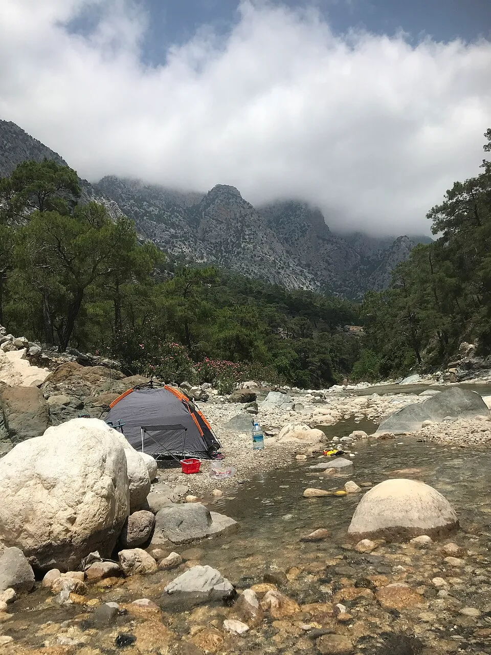 Geniş kayalıklarla çevrili berrak bir nehir yatağının kenarında kurulmuş gri ve turuncu bir çadır, arka planda yemyeşil çam ağaçları ve bulutların sardığı yüksek dağlarla, Antalya'nın Çitdibi bölgesindeki tırmanış maceraları için ideal bir kamp noktasını gözler önüne seriyor.