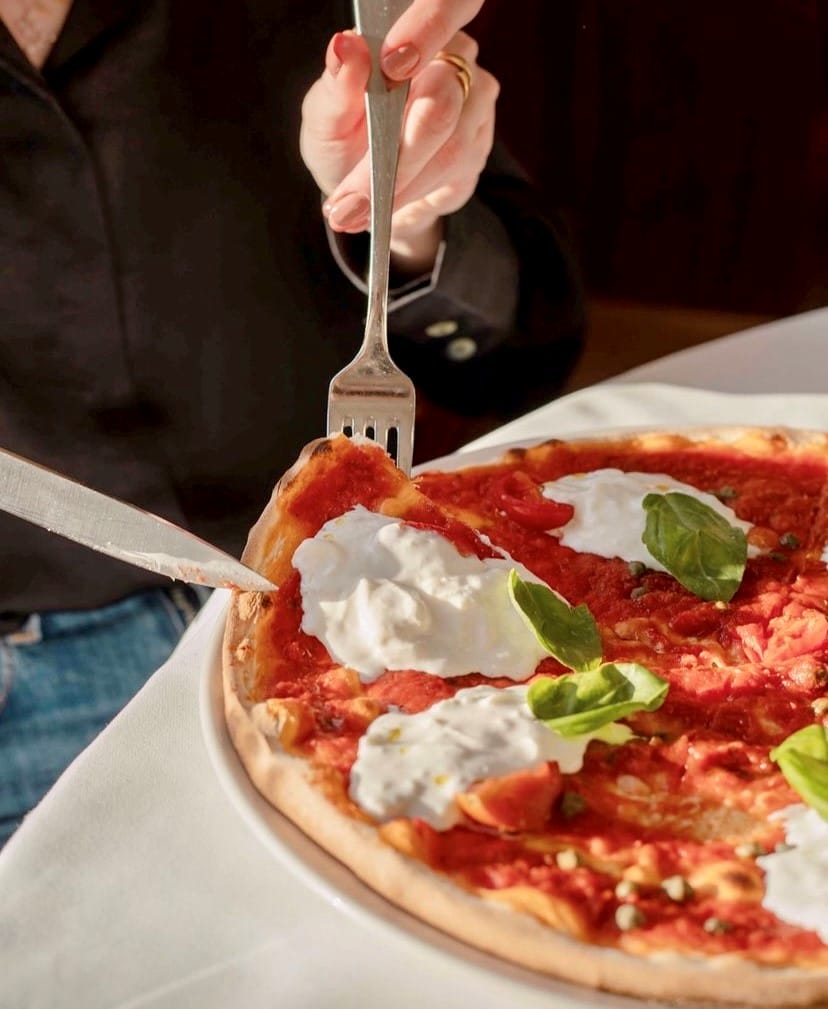 Etiler'deki Da Mario gibi seçkin bir mekanda, güneş ışığı alan beyaz masa üzerinde taze burrata peyniri, domates sosu ve fesleğen yapraklarıyla süslenmiş İtalyan pizzasının, ojeli bir el tarafından çatal ve bıçakla kesilerek servis edilmeye hazırlandığı iştah açıcı bir yakın plan.