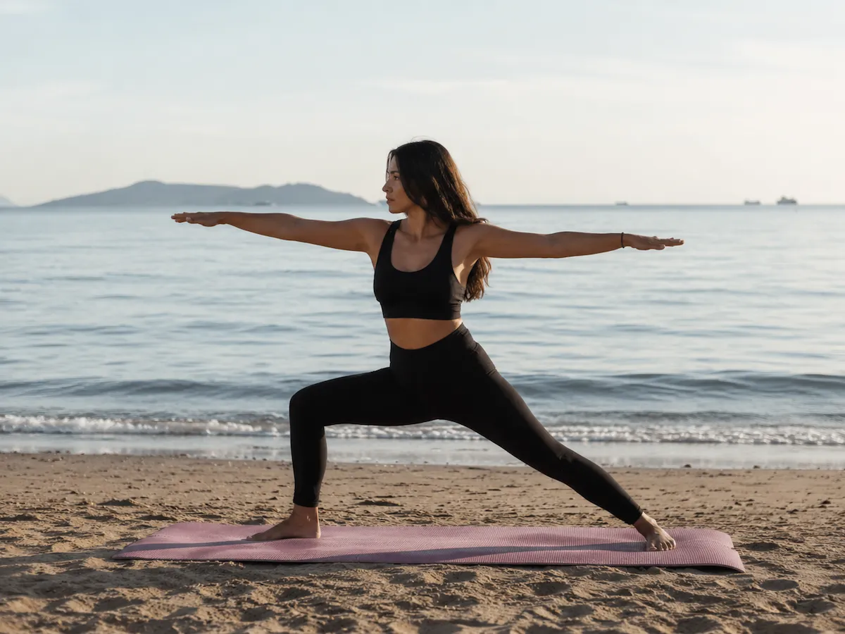 Kumlu bir kumsalda, sakin denizin kenarında, pembe bir yoga matının üzerinde savaşçı pozisyonunda yoga yapan uzun saçlı genç bir kadın, huzurlu bir anı yansıtırken, deniz kenarında yoga ve rahatlama deneyimini vurguluyor.