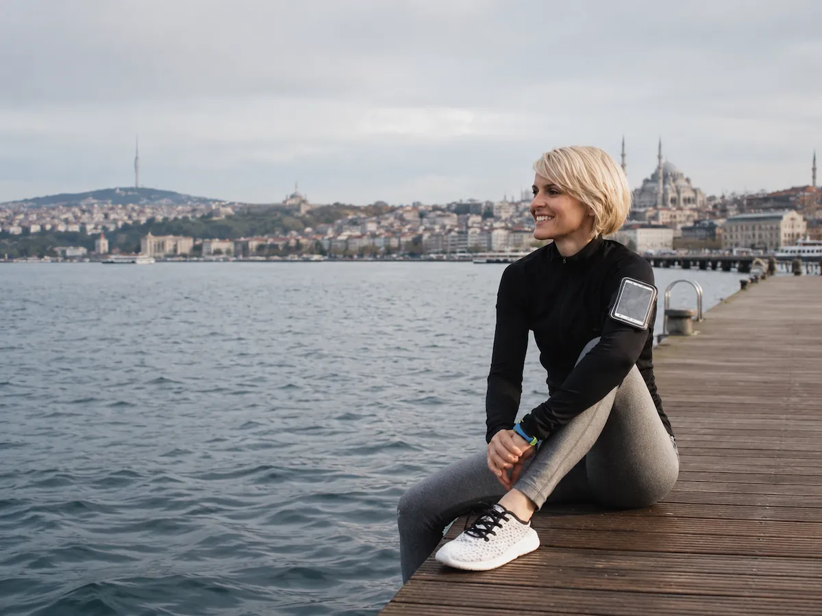 Spor giyimli, gülümseyen sarışın bir kadın, İstanbul silüetindeki cami ve kule manzarasına bakarak deniz kenarındaki ahşap iskelede oturuyor, yürüyüş ve hareket sonrası keyifli bir dinlenme anını yansıtıyor.