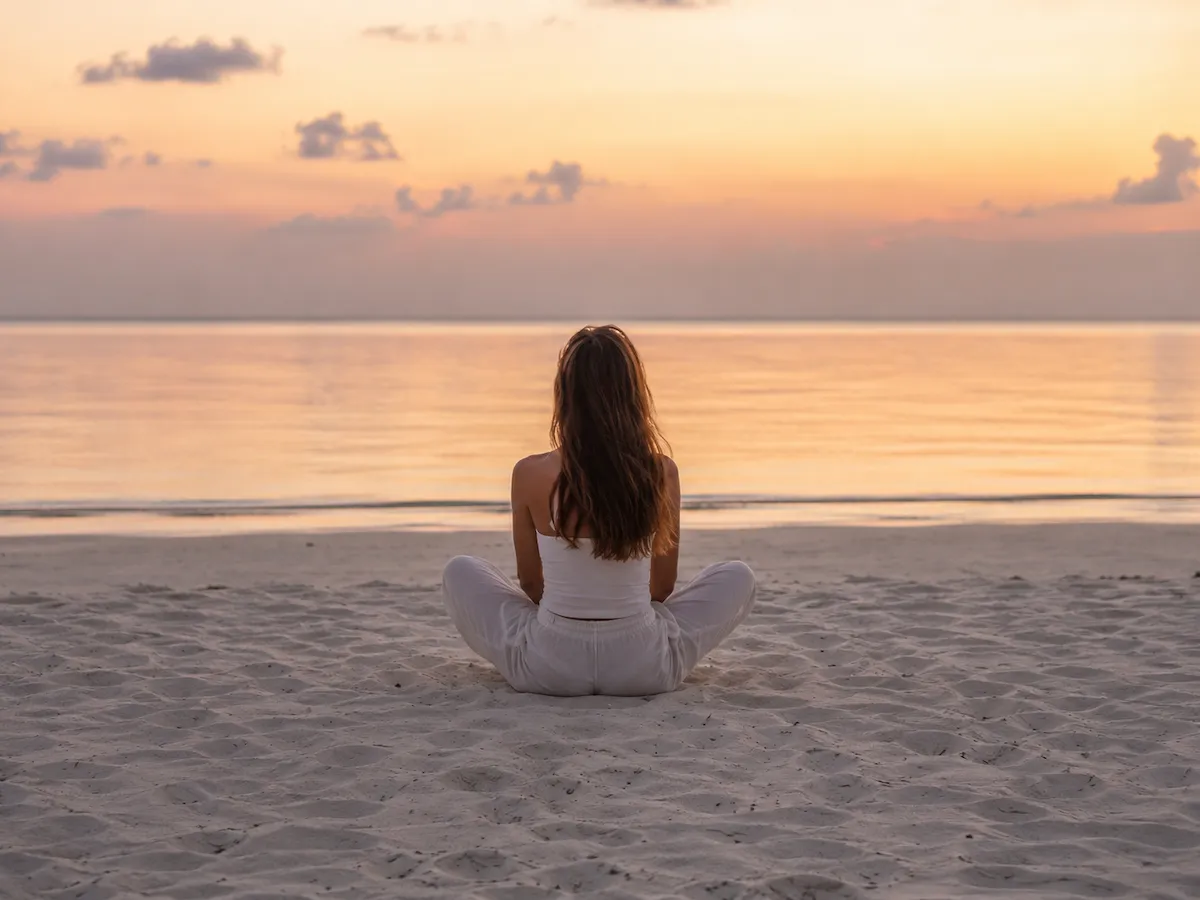 Gün batımının huzurlu renkleriyle aydınlanan sakin bir denizin kenarında, beyaz kıyafetler içinde kumsala bağdaş kurmuş genç bir kadın, deniz kenarında meditasyon yaparak zihinsel faydalar ararken arkadan görüntüleniyor, ruhsal dinginliği yansıtıyor.