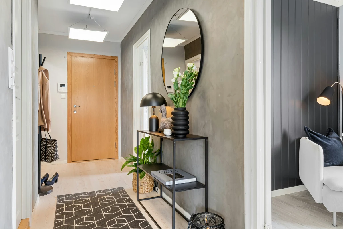 Açık renk ahşap parke zeminde siyah beyaz geometrik desenli bir halının uzandığı modern antrede, gri duvara monte edilmiş büyük oval aynanın altında siyah metal konsol üzerinde beyaz çiçekli bir vazo, siyah bir masa lambası ve dekoratif objeler dikkat çekerken, antrenin sonunda ahşap bir kapı ve sol tarafta askılıkla asılı bir mont görülmektedir.