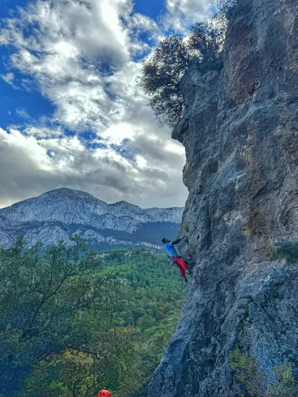 Mavi tişörtlü ve kırmızı pantolonlu bir dağcı, Geyikbayırı'nın sarp ve etkileyici kaya duvarında tırmanışını sürdürürken, arka planda yemyeşil ormanlar, heybetli dağlar ve bulutlu bir gökyüzüyle doğanın muhteşem manzarası uzanıyor.