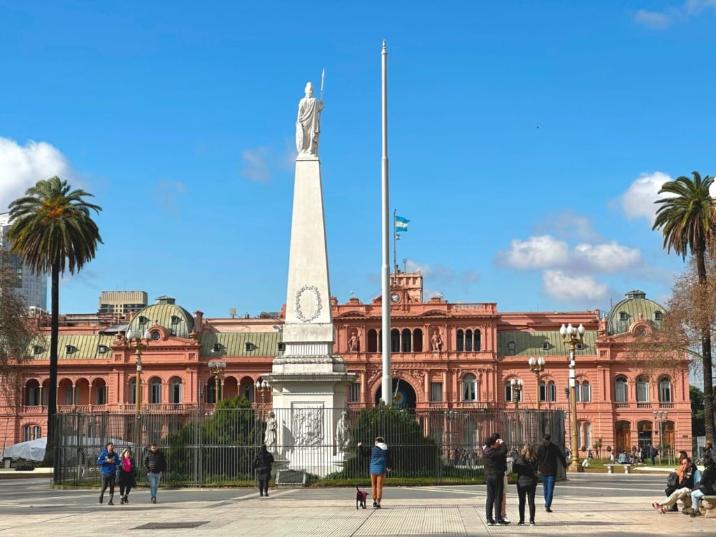 Buenos Aires'in siyasi tarihinin kalbi olan Plaza de Mayo'da, pembe renkli ve ikonik başkanlık sarayı Casa Rosada'nın önünde, beyaz bir obelisk anıt ve dalgalanan Arjantin bayrağı gökyüzüne uzanırken, insanların yürüyüş yaptığı ve dinlendiği, şehrin kültürel zenginliğini yansıtan hareketli ve geniş bir meydan manzarası.