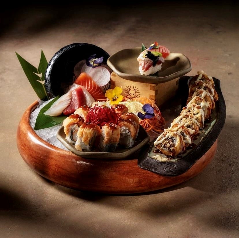 Etiler'in popüler mekanlarından Inari Sushi Omakase'den bir sunumda, ahşap bir servis tabağı üzerinde sergilenen somon ve çeşitli beyaz balık sashimi dilimleri, narince hazırlanmış susamlı ve kırmızı garnitürlü sushi roll'lar, özel soslarla kaplanmış çıtır roll'lar ve minik bir ahşap kutu üzerinde duran nigiri benzeri özenli bir parça, renkli yenilebilir çiçek ve sebze garnitürleriyle Uzak Doğu mutfağının zarafetini vurguluyor.
