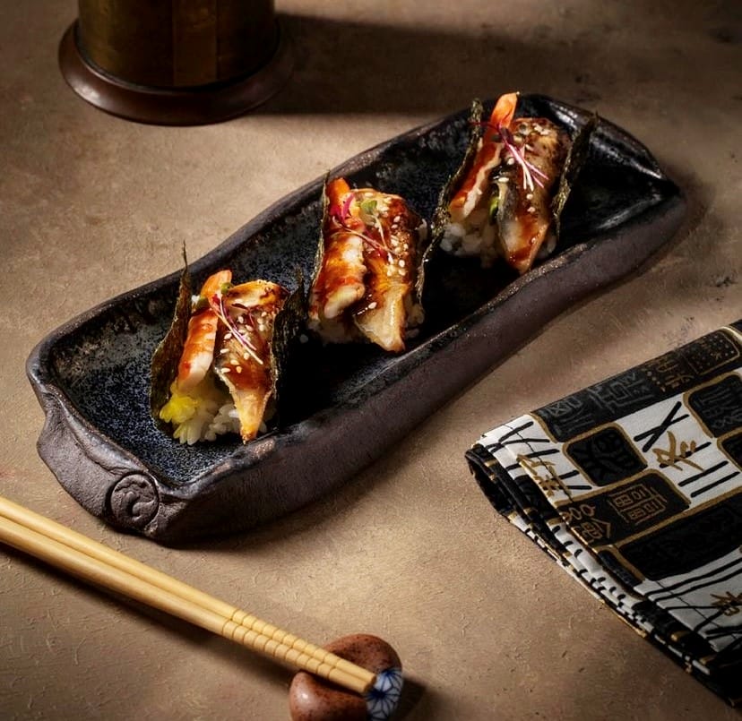 Etiler'deki en iyi mekanlardan Inari Sushi Omakase'nin Uzak Doğu mutfağı deneyimini yansıtan bir görselde, koyu renkli, dokulu bir tabakta parlak soslu ve susam taneleriyle süslenmiş üç adet ızgara balık ve karidesli suşi, yanında zarif yemek çubukları ve desenli bir peçeteyle iştah açıcı bir şekilde sunuluyor.