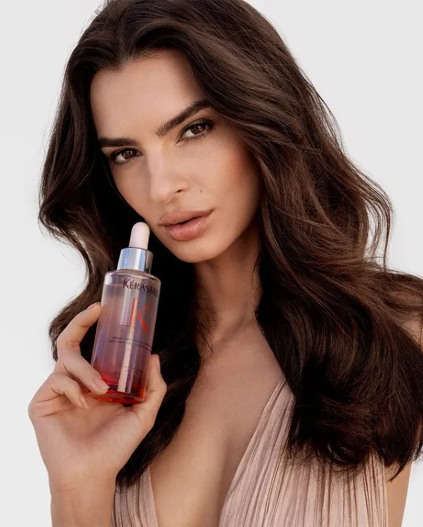 Uzun koyu dalgalı saçlara ve açık renk dekolteli bir elbiseye sahip model Emily Ratajkowski, beyaz bir fonda zarifçe poz verirken elinde Kérastase Genesis saç dökülmesine karşı güçlendirici serum şişesini dikkat çekici bir şekilde tutuyor.