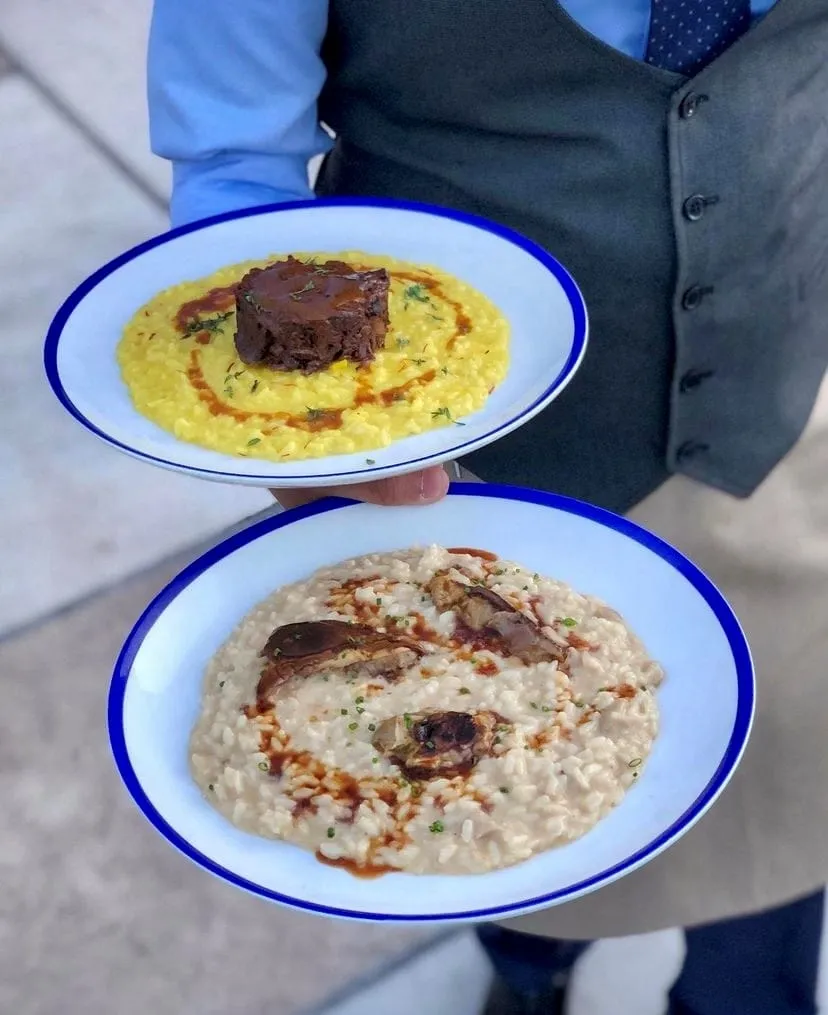 Bir garson, gri yeleği ve mavi gömleğiyle, biri sarı safranlı risotto üzerinde kahverengi et parçası, diğeri mantarlı kremalı risotto olan iki tabak İtalyan yemeği servis ediyor, her iki tabağın da sosları lezzetli görünüyor ve adeta la scarpetta yapmaya davet ediyor.