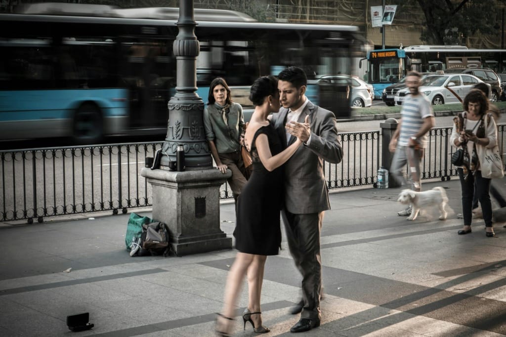 Buenos Aires sokaklarında tutkuyla tango yapan bir çift, şehrin dinamik atmosferi içinde akıcı hareketleriyle dans ederken, arkalarındaki bulanık şehir trafiği ve yakınlarındaki meraklı bir izleyici, kentin canlı ruhunu yansıtır.