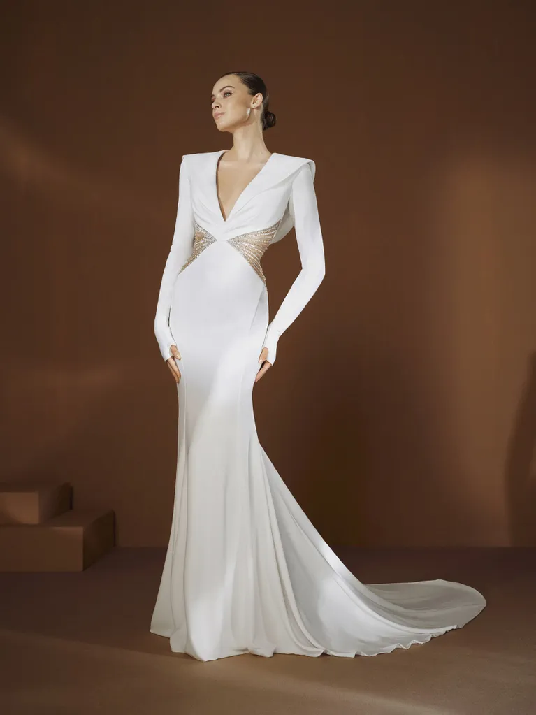 Pronovias tasarımı, derin V yaka ve transparan yan panellere sahip uzun kollu beyaz gelinlik modeli. Keskin omuz hatları ve zarif balık kesimiyle modern, güçlü ve iddialı bir siluet sunuyor.