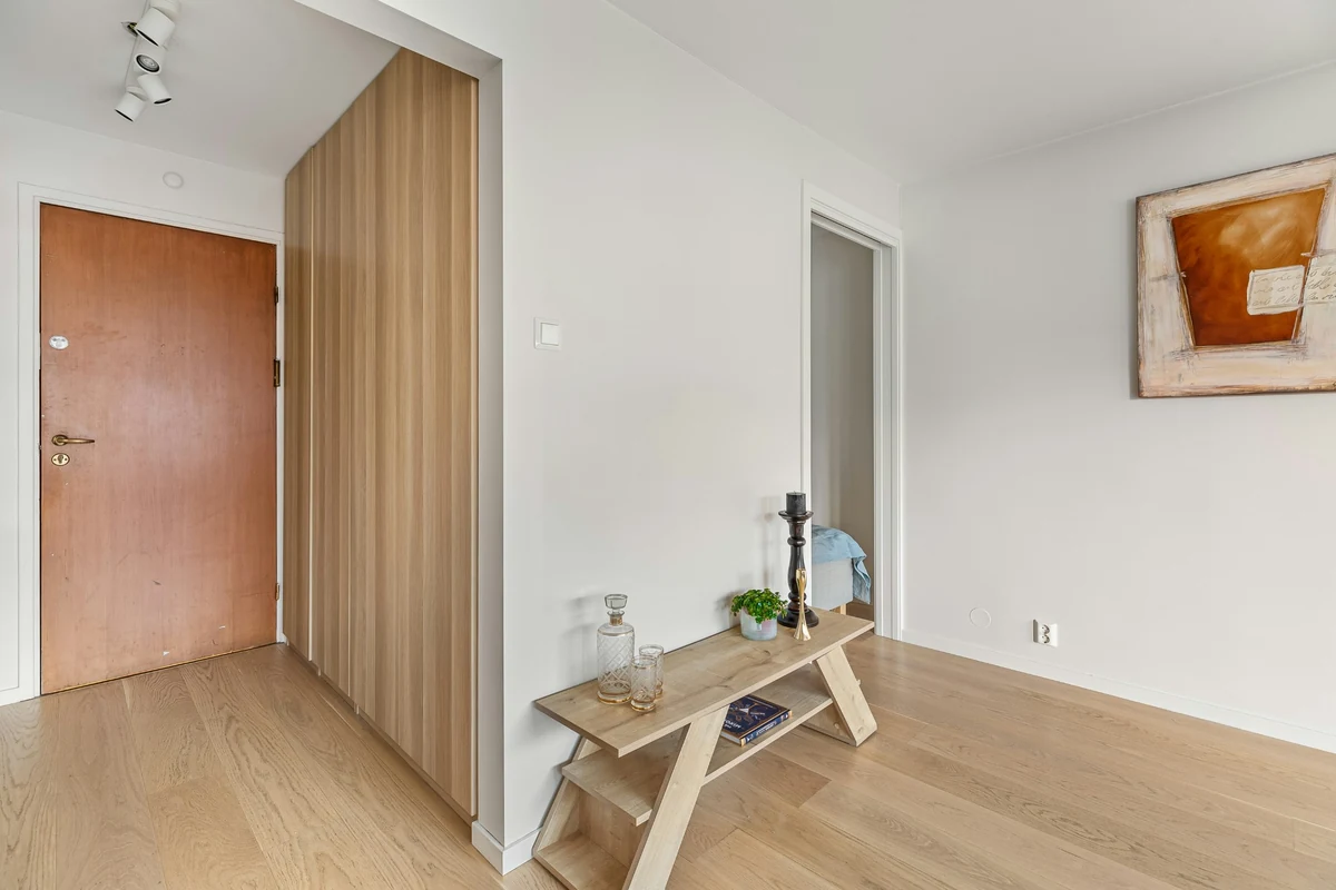Açık renk ahşap kapı, paneller ve parke zemini olan beyaz duvarlı sade bir antrede, üzerinde cam sürahi, bardaklar ve bitki bulunan ahşap konsol, ahşap detaylarla modern bir dekorasyon sergiliyor.