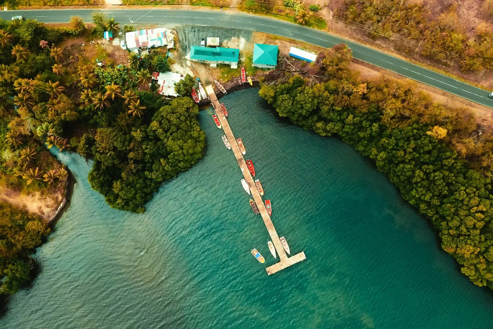 Saint Lucia'dan bir manzara sunan bu havadan çekilmiş fotoğrafta, turkuaz sulara doğru uzanan bir iskeleye demirlemiş çok sayıda renkli balıkçı teknesi, kıyı boyunca yemyeşil tropik bitki örtüsü ve yol kenarındaki küçük binalar belirgin bir şekilde göze çarpıyor.