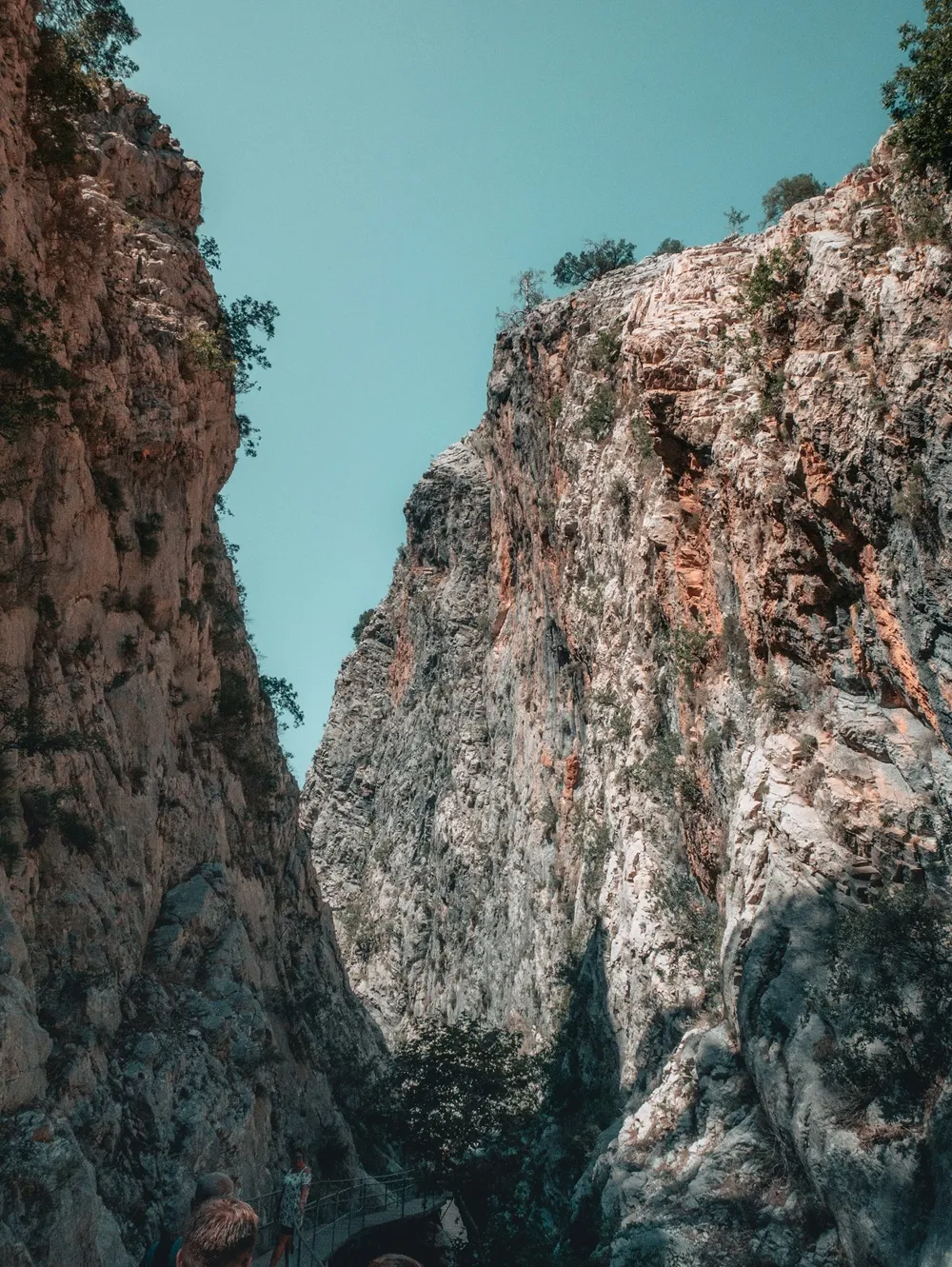 Sapadere Kanyonu'nun Antalya'da bulunan görkemli tırmanış rotaları sunan sarp kayalık yamaçları arasında, dar bir geçitten yükselen devasa taş duvarların ve alt kısımdaki yürüyüş yolunda gezinen ziyaretçilerin görüldüğü, mavi gökyüzüyle çevrili etkileyici bir doğa manzarası.