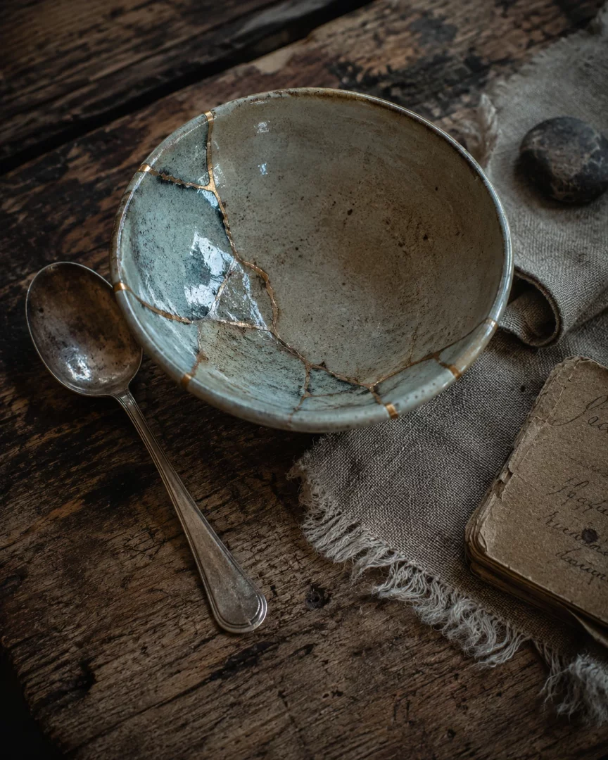 Koyu renkli rustik ahşap bir yüzey üzerinde, wabi-sabi estetiğini yansıtan, altın çizgilerle özenle onarılmış, mavi-yeşil tonlarında bir kintsugi seramik kase, yanında eski gümüş bir kaşık, dokulu çuval bezi, yuvarlak bir taş ve el yazması sayfalarla dolu yıpranmış bir defterle birlikte huzurlu bir kompozisyon oluşturuyor.