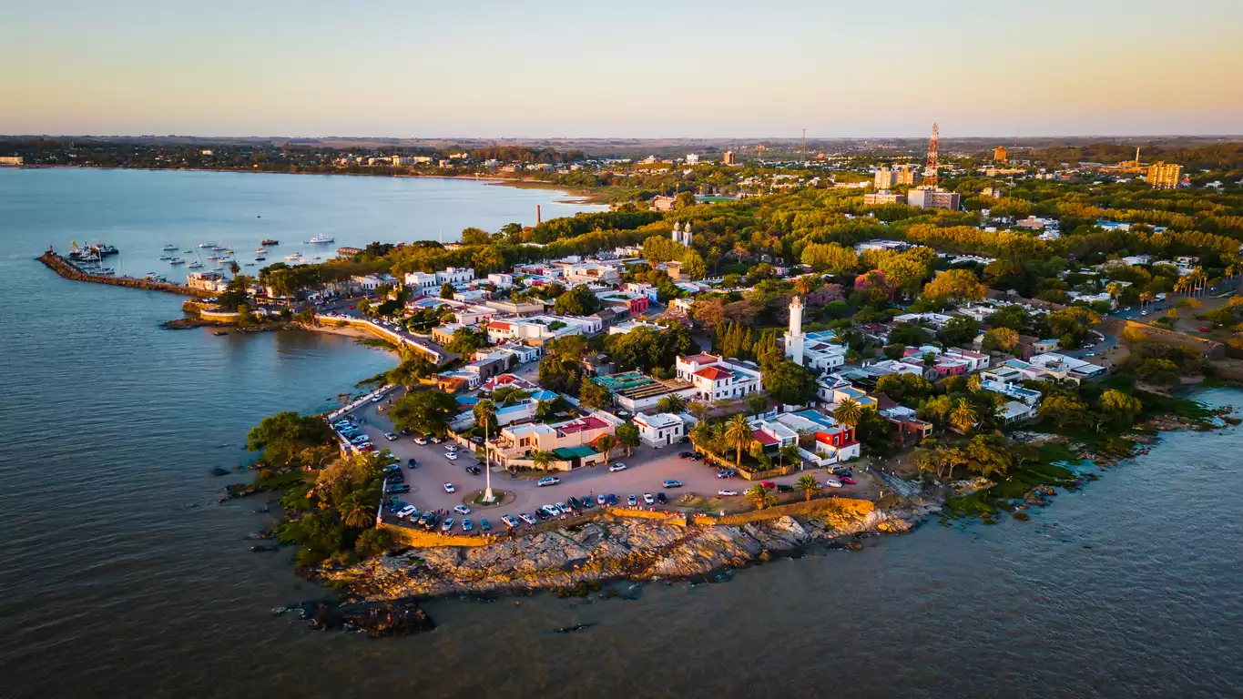 Buenos Aires'e yakın, UNESCO Dünya Mirası Listesi'ndeki tarihi Colonia del Sacramento şehrinin gün batımının yumuşak altın ışıkları altında, denize nazır park edilmiş arabalarla dolu dar sokaklarını, beyaz evlerini ve minik limanını gösteren büyüleyici kuşbakışı bir fotoğraf.