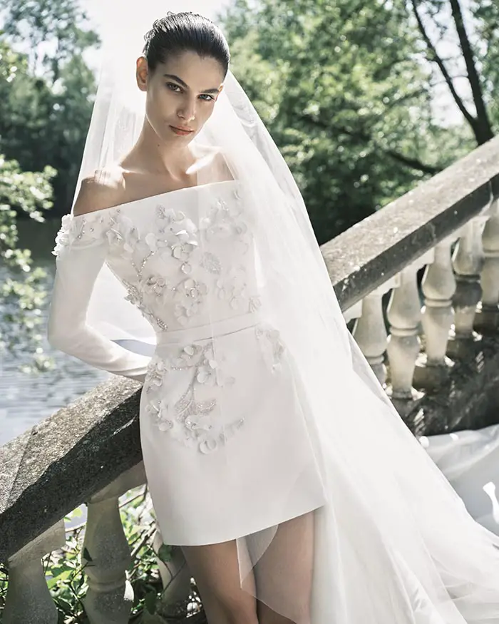 Elie Saab Gelinlik İlkbahar/Yaz 2025 koleksiyonundan, askısız, önü kısa arkası uzun kuyruklu, zarif çiçek ve boncuk işlemeli beyaz gelinlik giyen bir model, uzun tül duvağıyla taş bir korkuluğa yaslanmış, dış mekanda kameraya bakıyor.