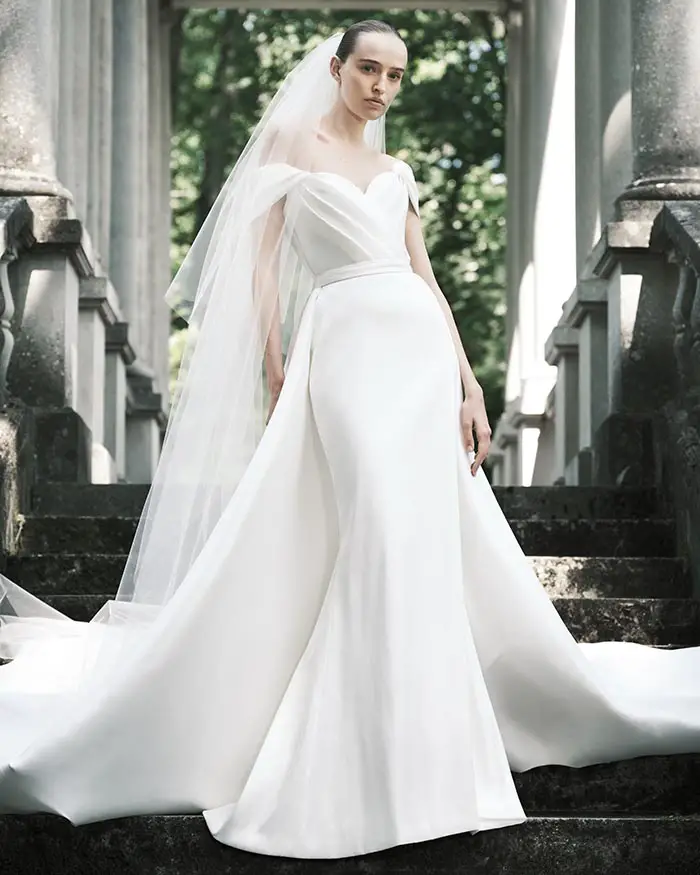 Bir model, Elie Saab BRIDAL İlkbahar/Yaz 2025 koleksiyonundan, dökümlü omuz dekolteli, kalp yakalı, uzun kuyruklu beyaz gelinliği ve şeffaf duvağıyla, eski taş sütunlu bir yapının merdivenlerinde duruyor.