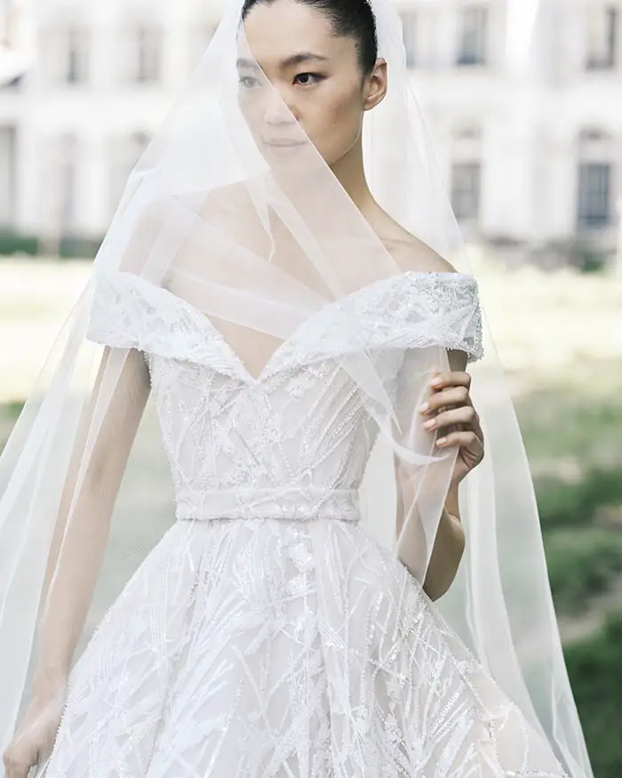 Elie Saab'ın İlkbahar/Yaz 2025 gelin koleksiyonundan bir görünüm; işlemeli, omuzları açık beyaz gelinliğin içindeki modelin yüzünün bir kısmı zarif tül duvakla örtülü, arka planda bulanık bir malikane ve yeşillik görülüyor.