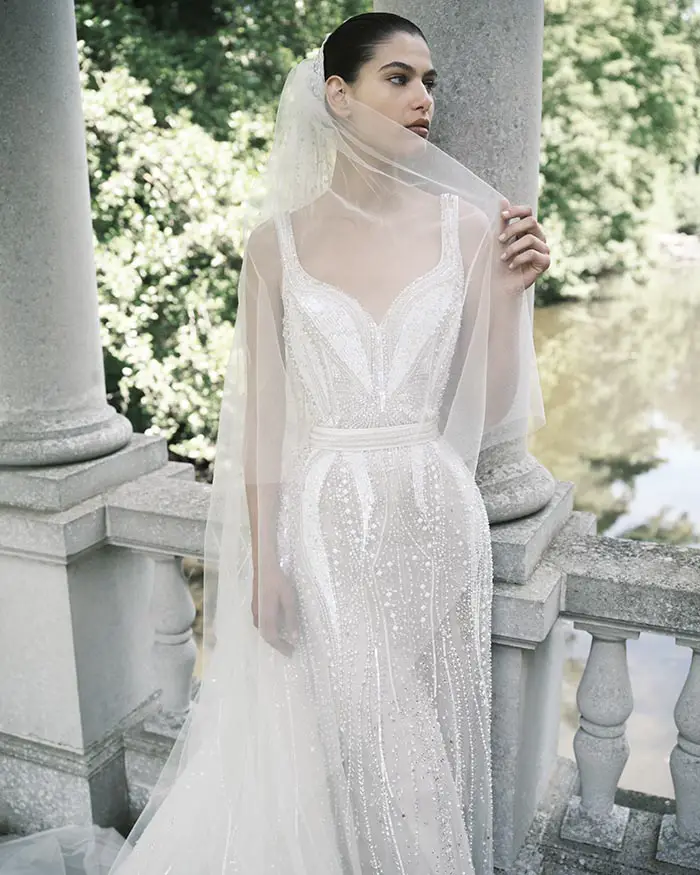 Elie Saab Gelinlik SS25 koleksiyonundan, askılı, derin kalp yaka ve incilerle yoğun işlemeli bembeyaz bir gelinlikle, taş bir balkonun trabzanında durup yüzünü örten tül duvağını hafifçe tutarak yemyeşil doğaya ve suya doğru zarifçe bakan bir gelin.