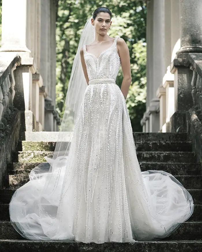 Elie Saab'ın İlkbahar/Yaz 2025 gelinlik koleksiyonundan bir modeli, baştan aşağıya pırıltılı inci ve boncuk işlemeli askılı, kalp yakalı zarif bir gelinlik ve uzun bir duvakla, yosun tutmuş eski taş merdivenlerde, kemerli sütunların ve yeşil ağaçların ortasında dururken gösteriyor.