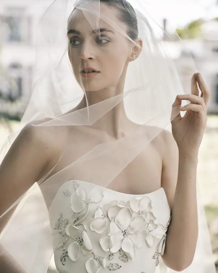 Elie Saab İlkbahar/Yaz 2025 gelinlik koleksiyonundan, omuzları açık, üç boyutlu çiçek işlemeleri ve gümüş detaylarla süslü beyaz bir gelinlik giyen, hafifçe yana dönük, düşünceli bir ifadeyle bakışlarını uzaklara yöneltmiş, yüzünü kısmen örten tül duvağıyla zarif bir gelin.