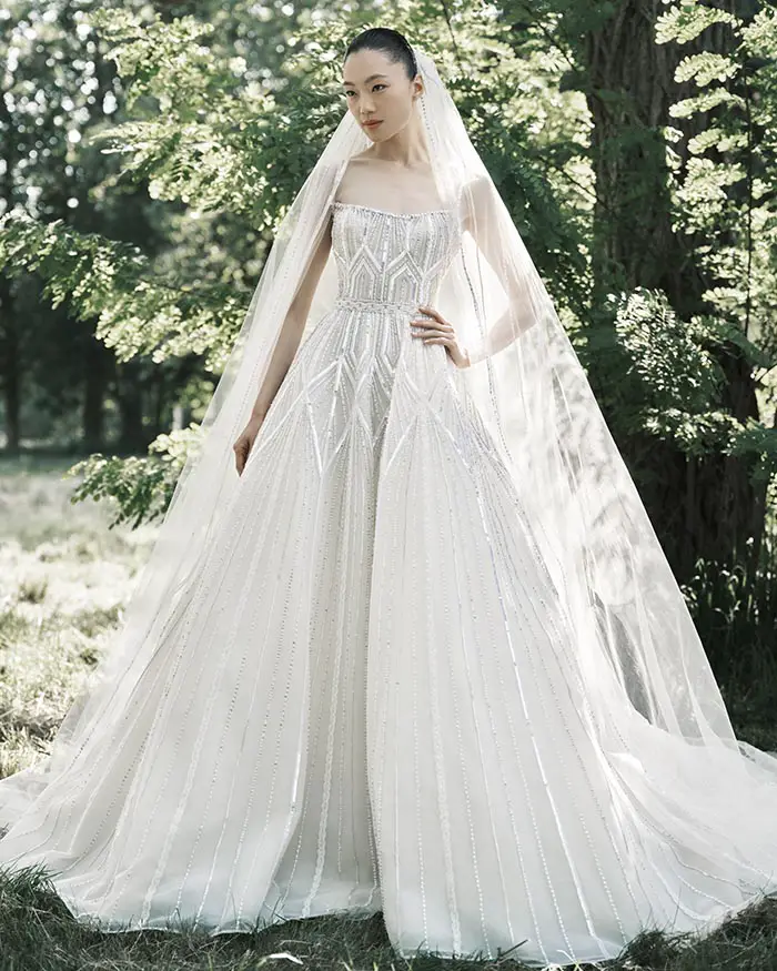 Doğal bir kır ortamında poz veren model, Elie Saab BRIDAL SS25 koleksiyonundan, straplez kesim, üzeri göz alıcı payet ve boncuk işlemelerle süslü, zarif bir gelinlikle ve uzun tülden bir duvakla asil bir duruş sergiliyor.