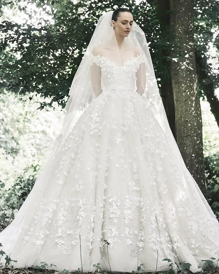Elie Saab İlkbahar/Yaz 2025 gelin koleksiyonundan beyaz, omuzları açık, üç boyutlu çiçek aplikeleriyle süslenmiş kabarık etekli bir gelinlik ve uzun duvak giyen model, yemyeşil ağaçların doğal fonunda zarif bir duruş sergiliyor.