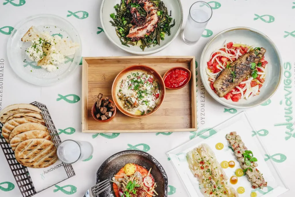 Selanik’in en iyi restoranları listesinde yer alan Glykanisos’un bereketli Ege deniz ürünlerini ve lezzetli mezelerini sergileyen bir masada, ızgara ahtapot, taze balık ceviche, pide ekmeği ve ouzo eşliğinde sunulan bol çeşitli bir gastronomi şöleni.