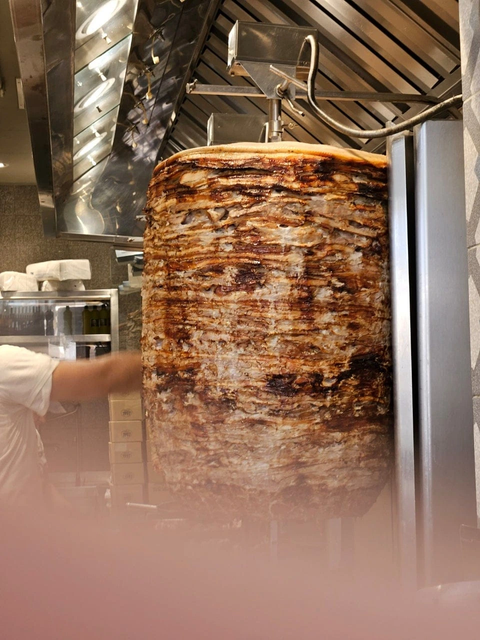 Pasparlak kızarmış et katmanlarından oluşan, büyük bir dikey döner şişinin parlak paslanmaz çelik yüzeylerin ve havalandırma davlumbazının altında bir mutfak ortamında durduğu, sol tarafta kısmen görünen bir çalışanın özenle hazırlandığı bu görüntü, Selanik'in en iyi sokak lezzetlerinden biri olan geleneksel Yunan döneri gyro'nun iştah açıcı görünümünü vurguluyor.