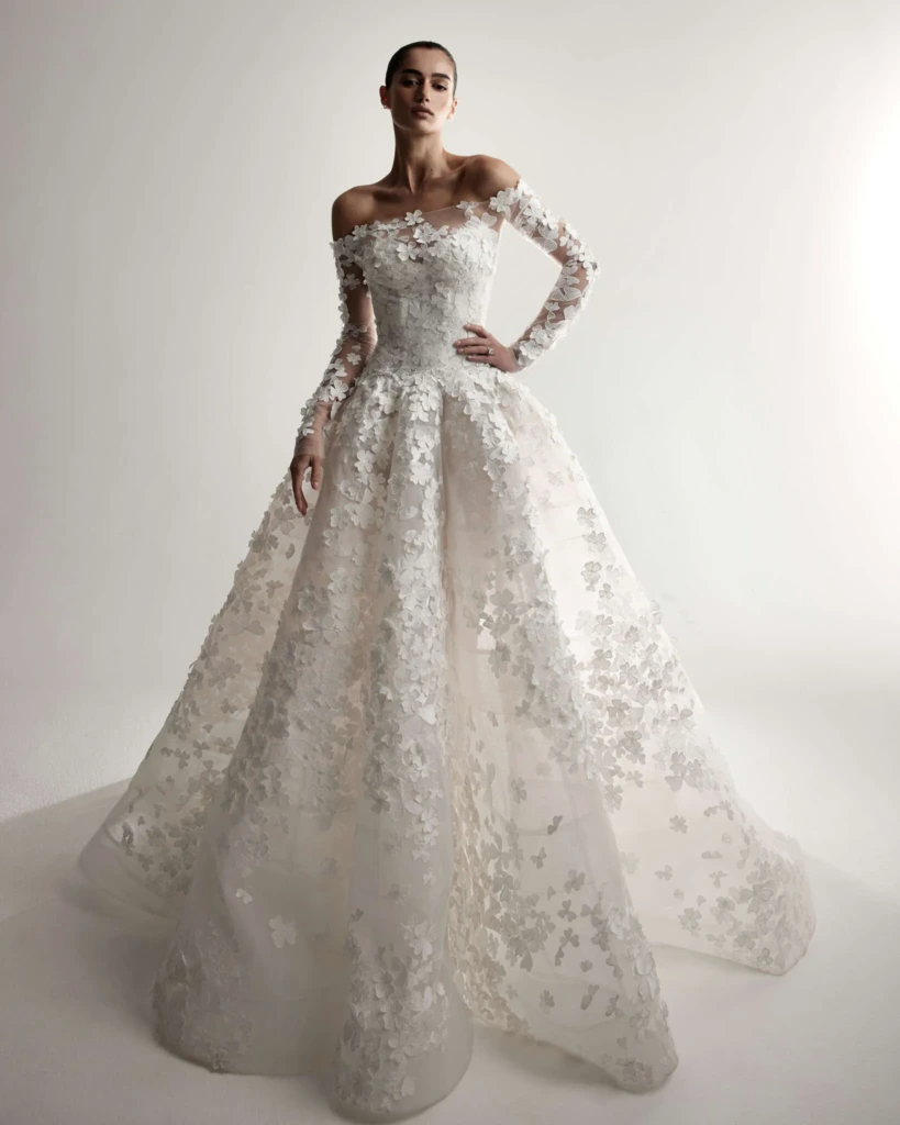 Monique Lhuillier 2026 koleksiyonundan, off-shoulder yakalı, uzun kollu ve üç boyutlu çiçek detaylı romantik ve ihtişamlı gelinlik modeli.