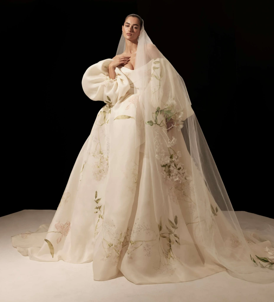 Monique Lhuillier 2026 koleksiyonundan, floral desenli, hacimli kollu ve uzun tül duvaklı dramatik ve sanatsal gelinlik modeli.
