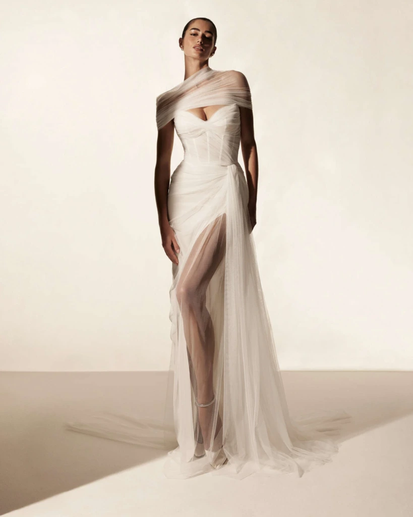Monique Lhuillier 2026 koleksiyonundan, tül drape detaylı, yırtmaçlı, heykelsi ve sofistike gelinlik modeli.