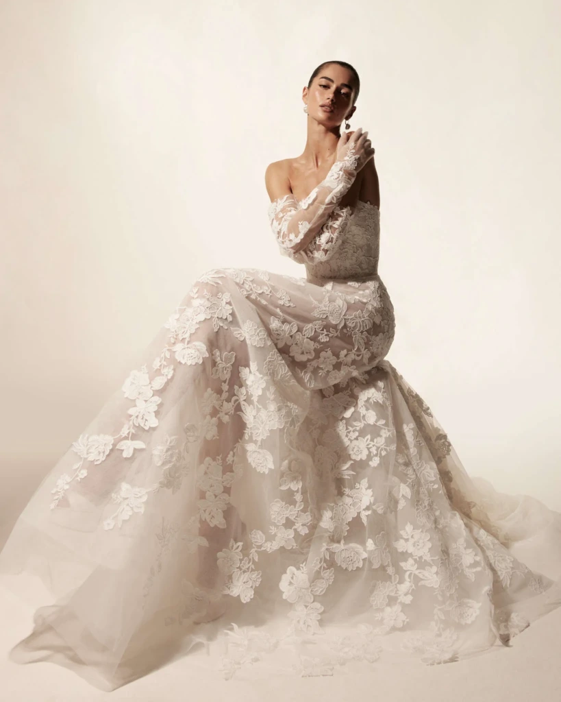 Monique Lhuillier 2026 koleksiyonundan, düşük omuzlu, uzun kollu ve tamamı dantel işlemeli zarif ve romantik gelinlik modeli.