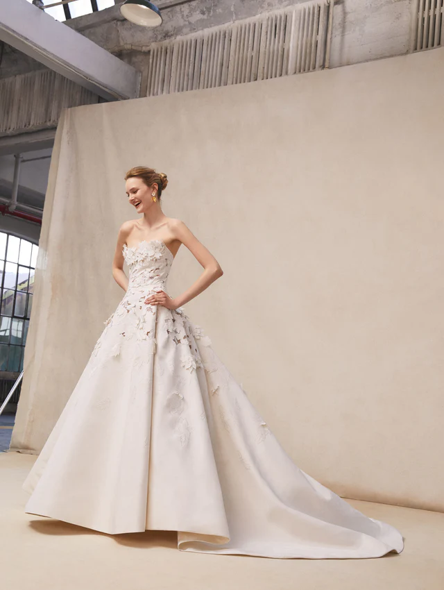 Oscar de la Renta 2026 koleksiyonundan, straplez yaka ve 3D çiçek detaylı A kesim saten gelinlik modeli.