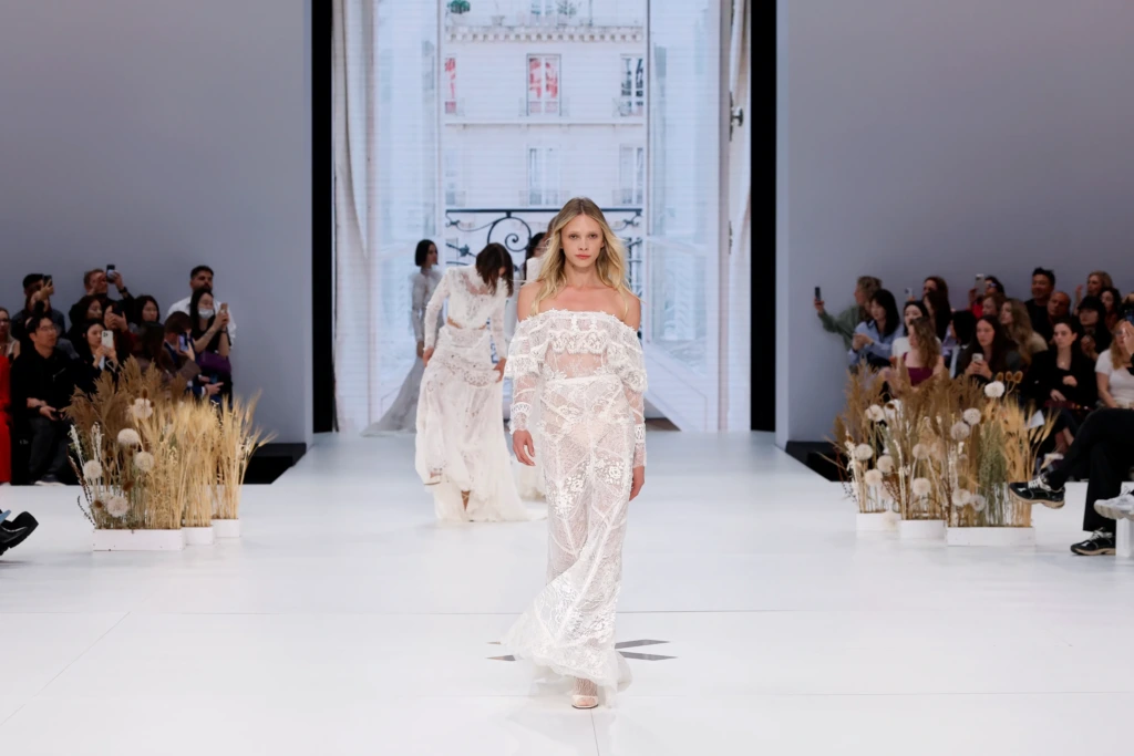 Barcelona Bridal Fashion Week'te podyuma çıkan model, Yolancris imzalı bohem gelinlik ile yürüyor. Transparan dantel dokular, düşük omuz kesimi ve doğal otlarla dekore edilmiş podyum detayı dikkat çekiyor.
