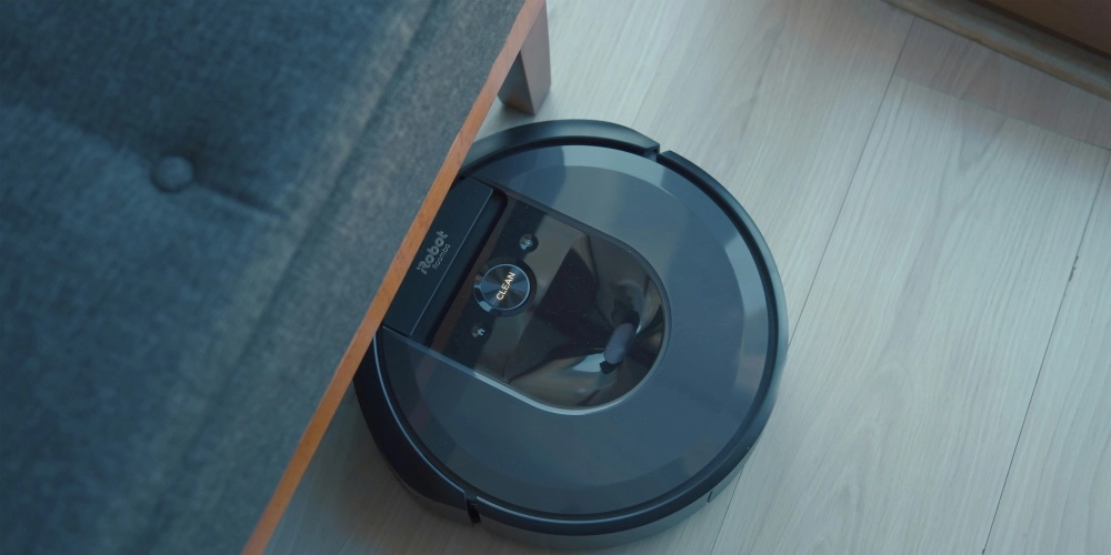 Koyu gri bir koltuğun ahşap bacağının altındaki açık renk parke zeminde, üzerinde Robot Roomba logosu ve CLEAN düğmesi bulunan koyu renk akıllı robot süpürge, mobilya altı temizliğini yaparken engel sensörlerinin etkinliğini gösteriyor.