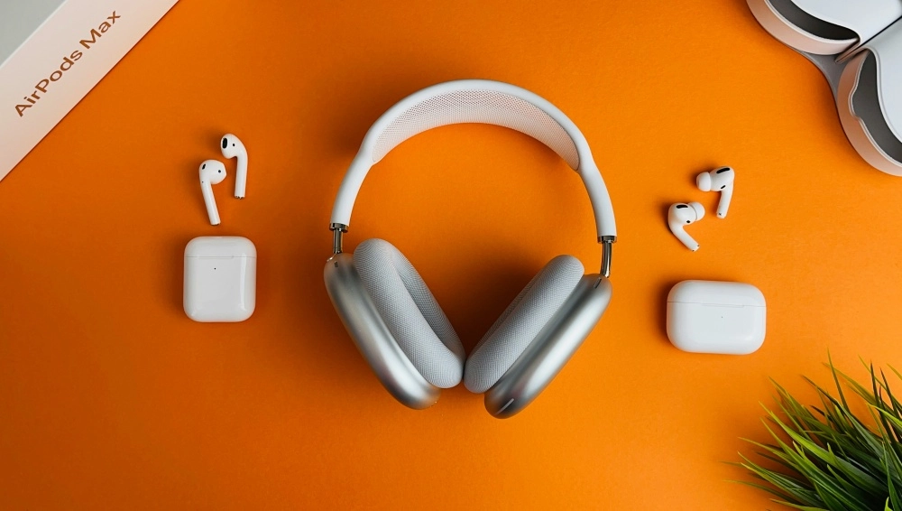 AirPods Max kulak üstü kulaklık ve AirPods Pro kulak içi modellerinin turuncu bir yüzeyde şarj kutuları ve diğer bir çift AirPods ile birlikte düzenli bir şekilde sergilendiği bir görünüm.