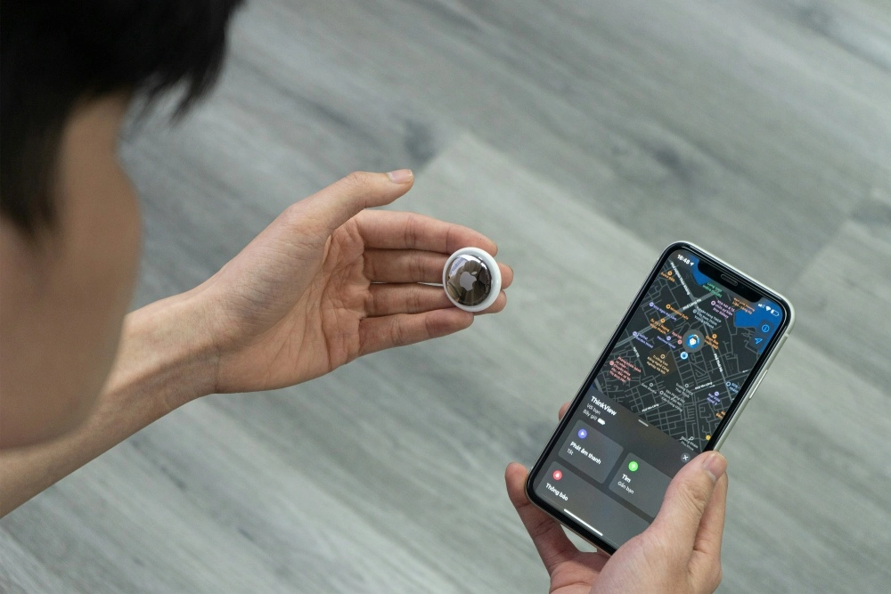 Bir kişi, beyaz Apple AirTag takip cihazını bir eliyle tutarken, diğer elindeki iPhone'un ekranında kayıp eşyaların yerini belirlemeye yarayan Find My (iPhone'umu Bul) uygulamasının harita görünümü ve ses çalma, yakınlarda bul gibi seçenekler açıkça görünüyor.