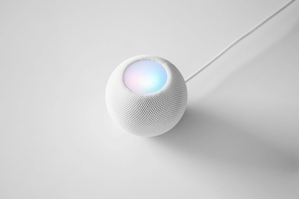 Minimalist beyaz bir zeminde, yuvarlak ve beyaz örgü kaplamalı Apple HomePod Mini akıllı ev hoparlörü, üst kısmında pastel tonlarda parlayan renkli ışıkları ve arkasından uzanan beyaz güç kablosuyla modern bir duruş sergiliyor.