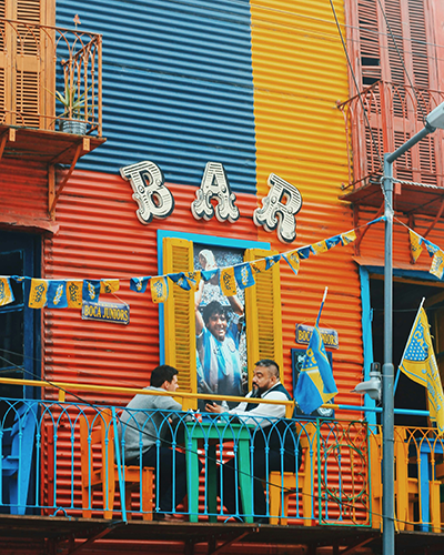 Buenos Aires'in ikonik La Boca mahallesindeki rengarenk, oluklu metal cepheli bir binanın BAR tabelalı üst kat balkonunda, sarı ve mavi Boca Juniors bayrakları arasında oturan iki adam, pencereden görünen milli takım forması giymiş bir figürle şehrin tango ve futbol tutkusunu canlı bir şekilde betimliyor.