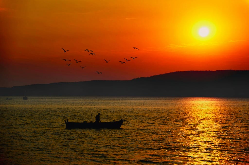 Çanakkale'nin dingin suları üzerinde batan güneşin turuncu ve altın tonlarındaki yansımaları, uzak ufukta siluet oluşturan tepeler ve gökyüzünde süzülen kuş sürüleriyle birlikte küçük bir teknedeki balıkçının huzur dolu anını yansıtırken, bölgenin doğal güzelliklerinin Çanakkale seyahat rehberindeki önemini vurguluyor.