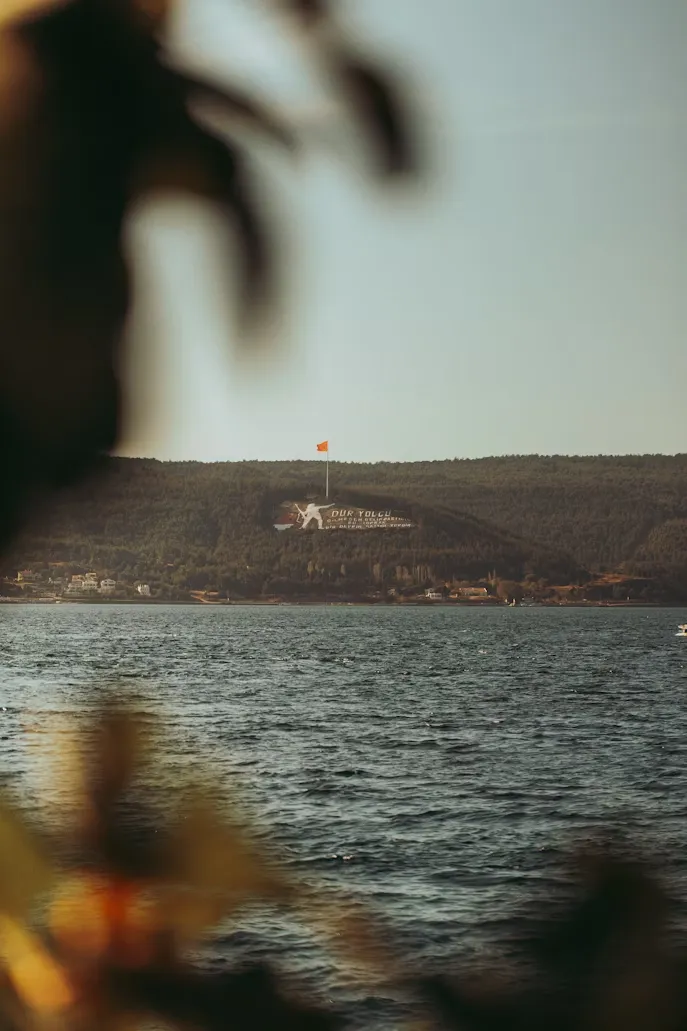 Ön planda bulanık yeşilliklerle hafifçe çevrelenmiş, Çanakkale Boğazı'nın hafif dalgalı sularının ötesinde, arka planda uzanan yemyeşil tepenin yamacında, büyük beyaz harflerle yazılmış Dur Yolcu anıtı ve üzerinde gururla dalgalanan parlak kırmızı Türk bayrağının net bir şekilde görüldüğü tarihi bir manzara.