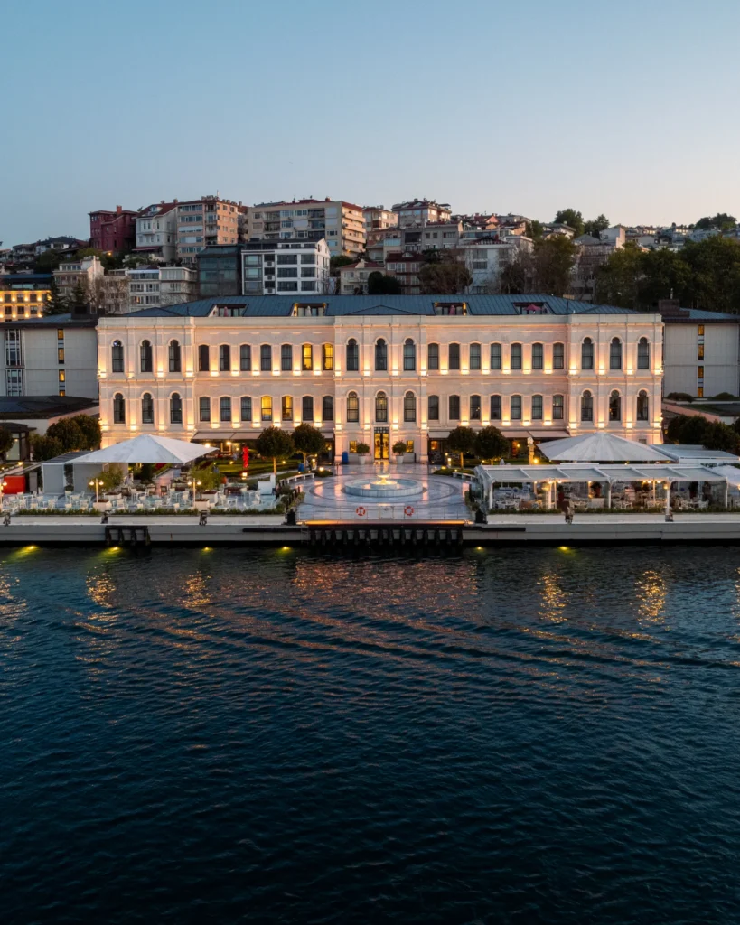 Akşam alacakaranlığında Boğaz kıyısında parıldayan, tarihi Çırağan Sarayı oteli pencereleri ve cephesi ışıklandırılmış halde, önündeki şık açık hava restoranları ve çeşmeli rıhtımıyla sakin sulara nazır bir manzara sunarken, arka planda tepeye sıralanmış İstanbul evleri gözüküyor.