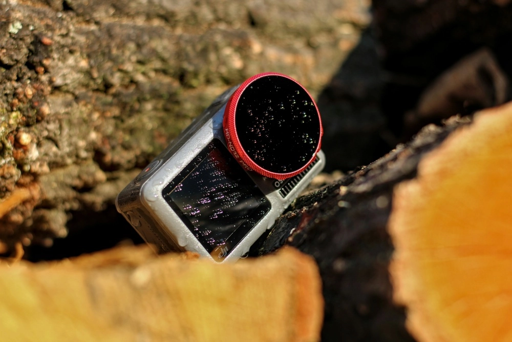 Bir aksiyon kamerası olan ve üzerinde HD-ND32 PRO yazan kırmızı kenarlı filtre lensi ile ön ekranında su damlacıkları bulunan gümüş renkli DJI Osmo Action, doğal bir ağacın dokulu yüzeyinde eğik durarak dış mekan kullanımına uygunluğunu vurguluyor.