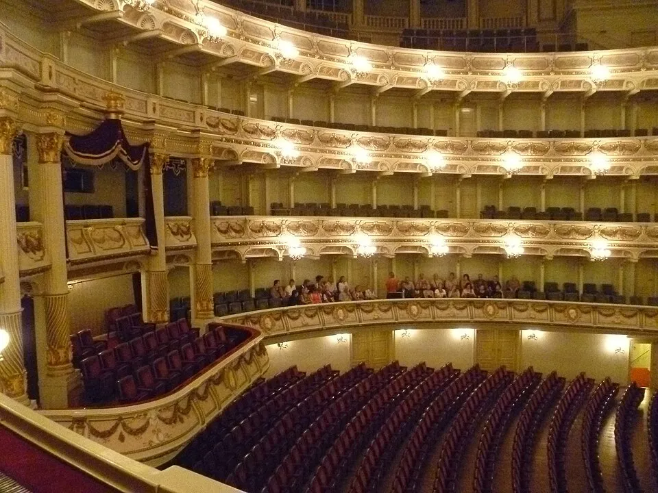 Dresden Semperoper'in altın rengi detaylarla bezenmiş, çok katlı balkon ve locallara sahip görkemli iç görünümünde, kadife kırmızı koltuklar sahneye doğru zarifçe sıralanırken, bazı balkon ve localarda oturan seyirciler bu tarihi opera binasının eşsiz mimarisini ve atmosferini hissetmektedir.
